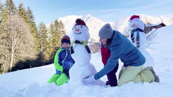 Mother and Two Kids Build Together Snowman in Mountains