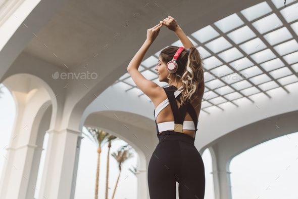 Portrait from back attractive sportswoman with long curly hair in headphones stretching hands above