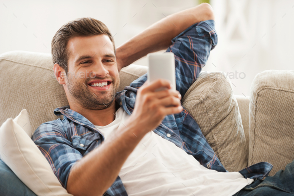 Staying in touch at home. Cheerful young man holding mobile phone and smiling while lying on sofa
