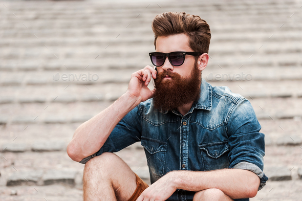Staying in touch. Confident young bearded man talking on the mobile phone while sitting outdoors