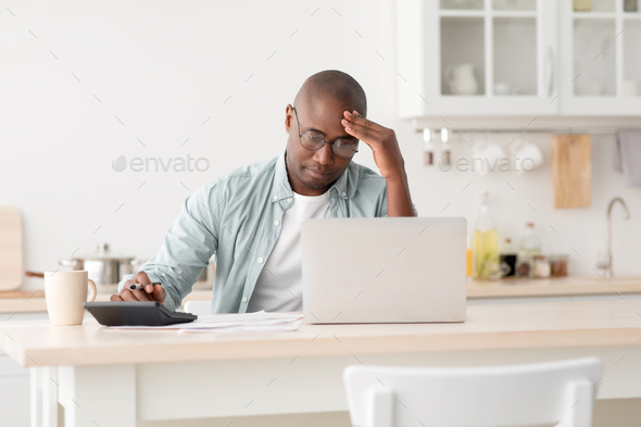 Stressed mature black man using calculator and laptop computer, calculating taxes, checking monthly