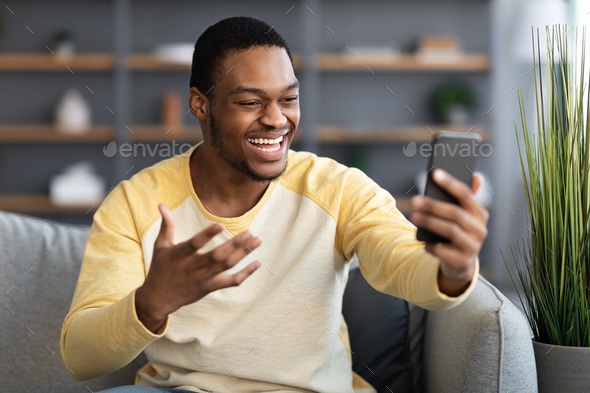 Joyful black man having video conversation on phone, home interior ...