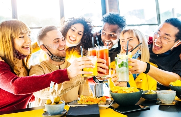 Happy multiracial friends wearing face masks toasting cocktails at bar restaurant
