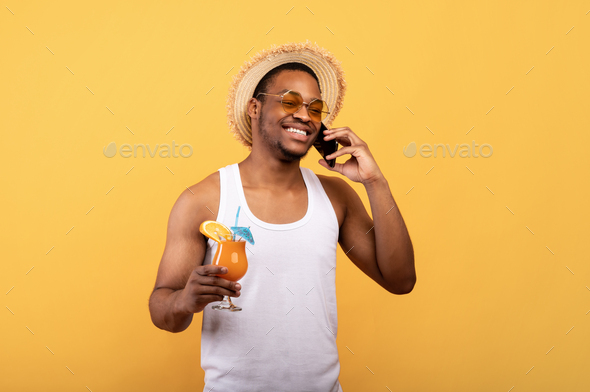 Positive black guy in trendy outfit holding tropical fruit cocktail and speaking on smartphone on