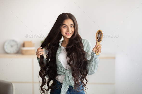 Portrait of happy Indian lady brushing her long dark hair with wooden brush at home