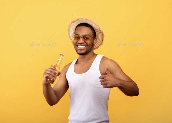 Handsome black guy in summer outfit holding beer bottle and showing thumb up gesture on yellow