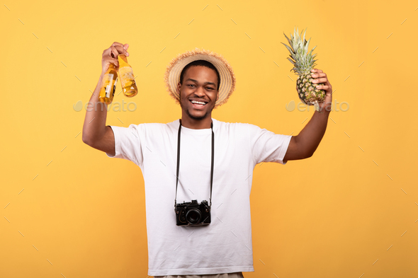 Portrait of handsome black guy in summer clothes with camera holding beer bottles and pineapple