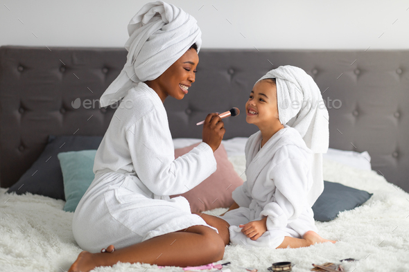 Happy black mom and her cute daughter wearing bath robes and towels, applying makeup on family spa