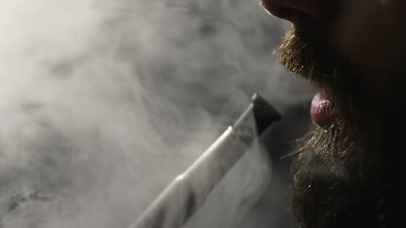 Young bearded man smokes a hookah and blow out smoke closeup on black background slowmotion