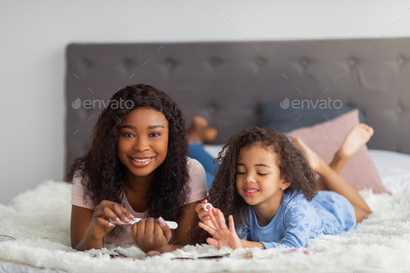 Joyful black mother and her little daughter doing manicure, lying on bed at home