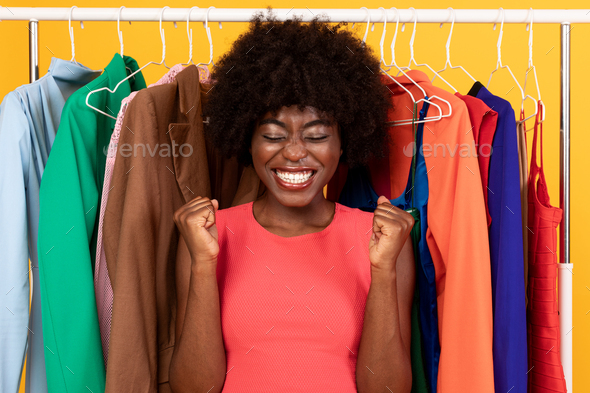 Excited Black Lady Shaking Fists Near Clothing Rail, Yellow Background