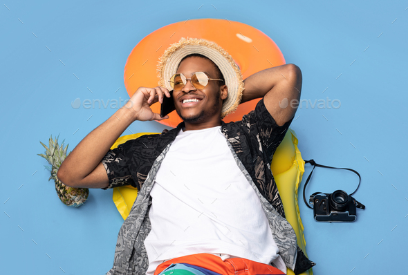 Funky black guy in trendy summer clothes lying on inflatable lilo, speaking on smartphone over blue