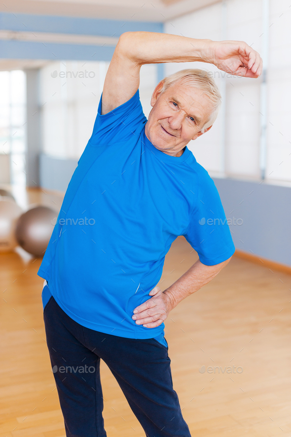 Staying active. Cheerful senior man doing stretching exercises and smiling while standing indoors