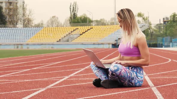 Girl Doing Online Workout with Laptop in Empty Stadium. Charming Blonde. During Quarantine.
