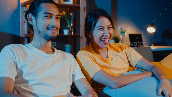 man and woman smile and laugh lay down on sofa in living room at night watch comedy movie.