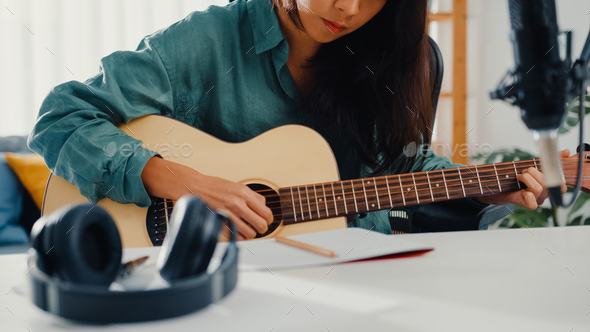 Asian woman songwriter play acoustic guitar listen song from smartphone think and write notes.