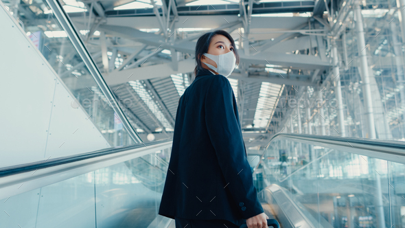 Asian business girl wear face mask drag luggage stand on escalator look around.