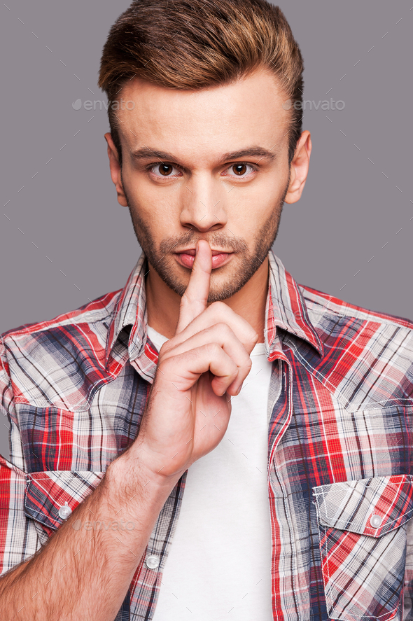 Silence please! Young man giving silence gesture against grey background