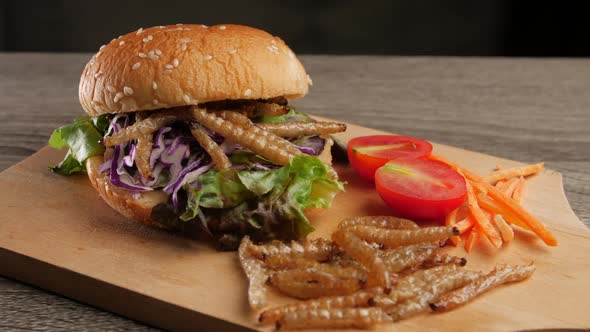 Burger with fried bamboo caterpillar and vegetable salad on wooden plate. alt