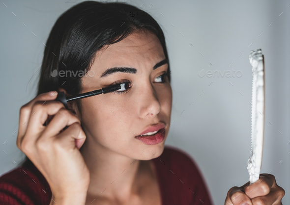 Beautiful young woman putting on mascara make up while looking in the mirror