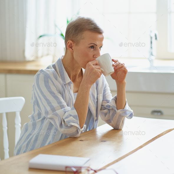 Attractive mature woman starting day with coffee while sitting at wooden table in kitchen - Stock Photo - Images