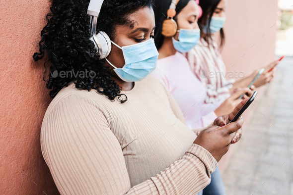 Young multiracial friends wearing protective masks while using mobile phone outdoors