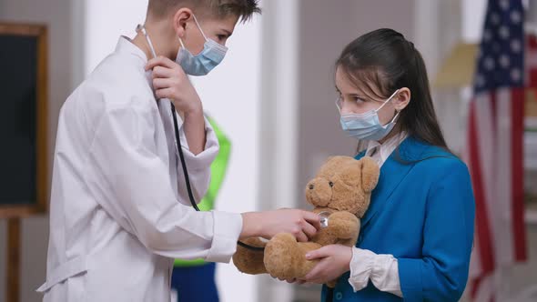 Focused Little Doctor in Coronavirus Face Mask Listening Teddy Bear with Stethoscope alt
