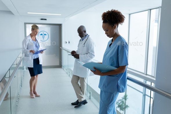 Three diverse male and female doctors standing in hospital corridor looking at medical documentation