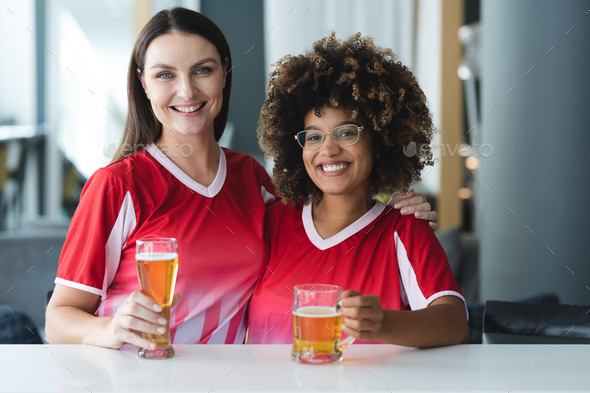Portrait of two diverse female sports fans having beer smiling at bar