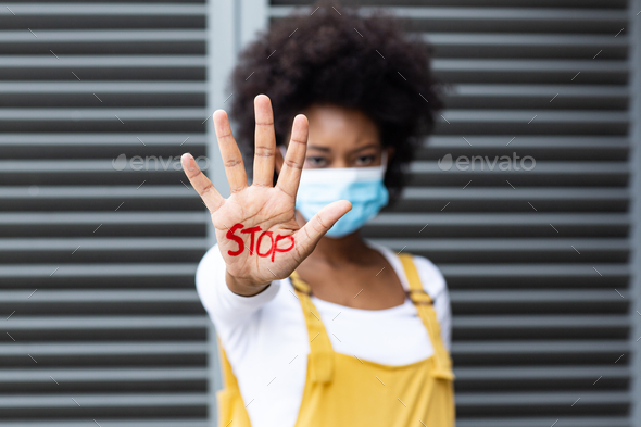 Portrait of mixed race woman wearing face mask making stop gesture with hand covered in writing