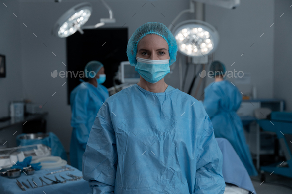 Portrait of caucasian female surgeon wearing face mask, surgical cap and gown in operating theatre - Stock Photo - Images