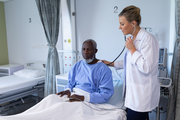 Caucasian female doctor examining with stethoscope african american male patient in hospital room - Stock Photo - Images