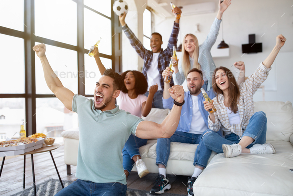 Group of young multiracial friends watching soccer game on TV with snacks and beer, cheering for