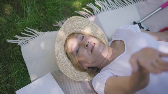 Top View Happy Senior Woman in Straw Hat Lying on Blanket in Park Looking at Sunshine Through alt