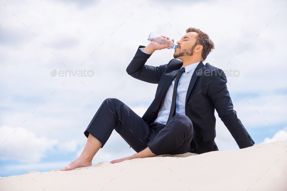 Refreshing his mind. Young businessman drinking water while sitting on the top of sand dune