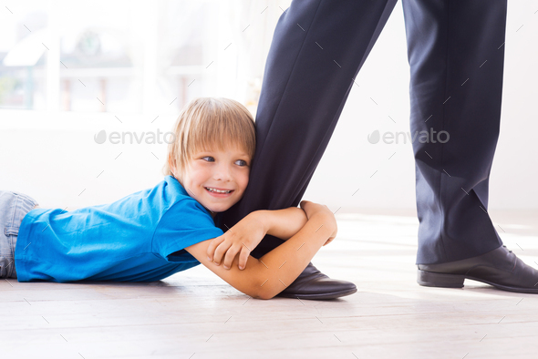 Stay with me! Playful little boy embracing his father leg and smiling while lying on the floor