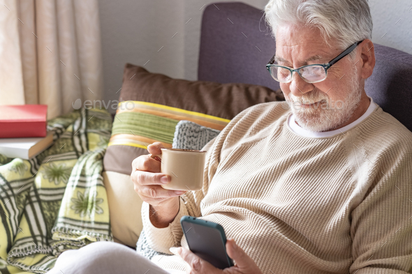 Handsome white-haired elderly man in lockdown at home using his smart phone while drinking