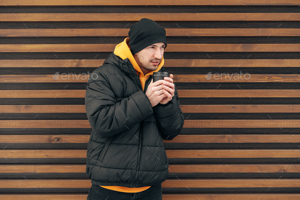 Young man in winter clothes standing with coffee to go in a street