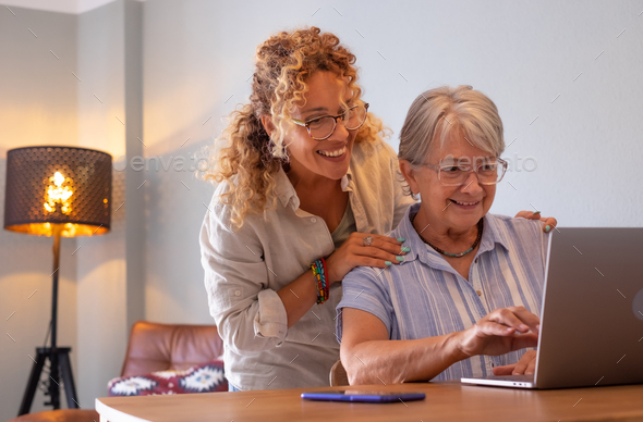Beautiful blonde adult daughter teaching her mother how to use the network with digital laptop - Stock Photo - Images
