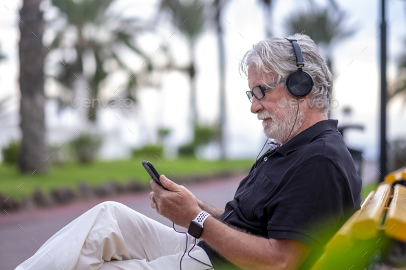 Portrait of senior white-haired man sitting in a bench and listening to music with headphones - Stock Photo - Images