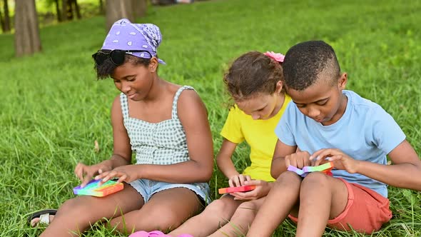 Children of Different Races Sit on the Grass and Play with an Antistress Toy Popit alt