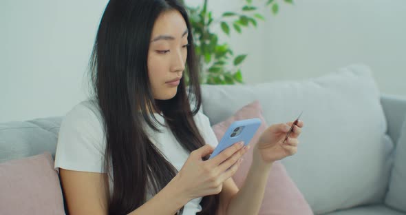 Beautiful Asian Woman Sitting on Couch Holding Cellphone Using Credit Card Makes Instant Payment alt