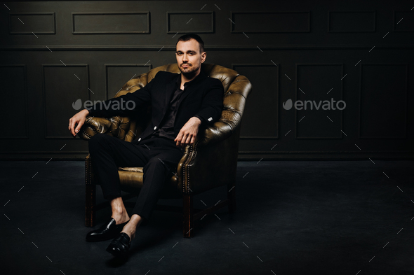 A male businessman in a black suit sits relaxed in a chair against a dark  background Stock Photo by Lobachad