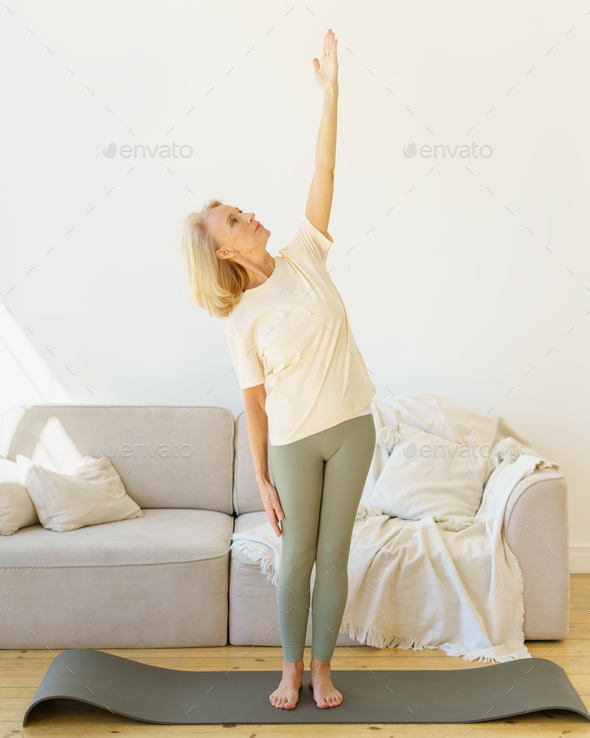 Healthy mature woman standing in yoga pose while exercising in living room at home - Stock Photo - Images