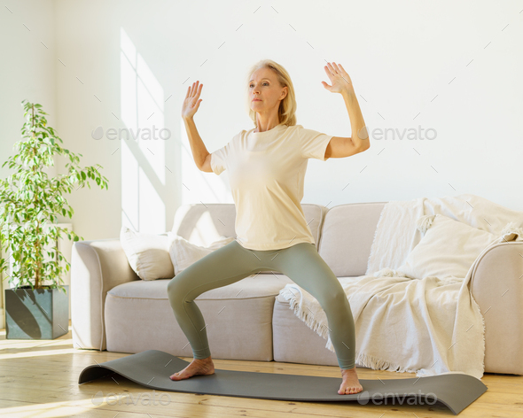 Focused retired mature woman in sportswear practicing yoga asana at home while standing on mat - Stock Photo - Images