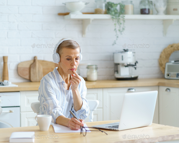 Focused mature woman in headphones sitting in kitchen and making notes, studying online on laptop - Stock Photo - Images
