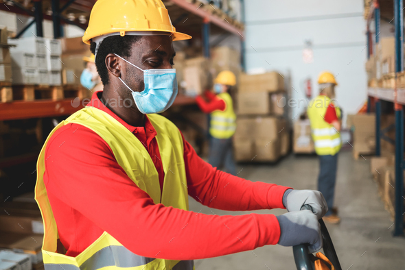 Afro american worker inside warehouse pulling a pallet truck while wearing safety mask - Stock Photo - Images