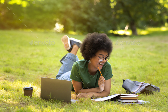 Cheerful black student girl doing homework with laptop computer outdoors
