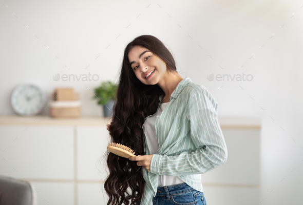 Portrait of lovely Indian lady brushing her wavy long hair, using wooden brush. Domestic spa salon