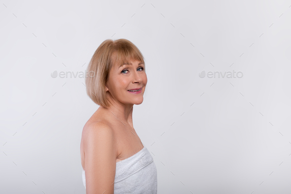 Anti-aging care. Attractive senior woman in towel pampering her sensitive mature skin on light
